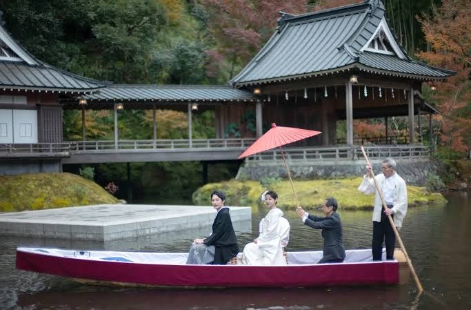 A bride and groom in traditional Japanese wedding kimono crossing Asaba's pond by boat under a red parasol